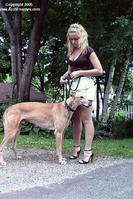 Woman standing next to a dog on a leash in an outdoor setting with trees and a house in the background in high heels