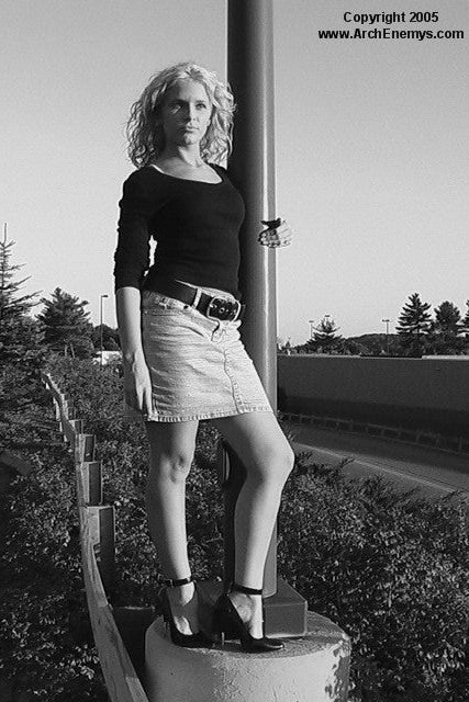 Black and white photo of a woman standing outdoors with trees and a building in the background in high heels