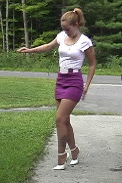 Woman in a white top and purple skirt standing on a sidewalk next to a red car.