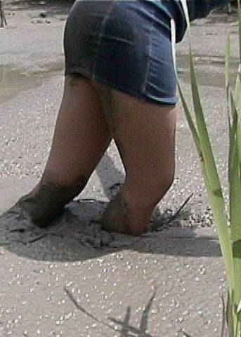 Person wearing a blue skirt and sandals with mud on feet, standing on sandy ground with grass.
