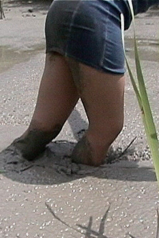 Person wearing a blue skirt and sandals with mud on feet, standing on sandy ground with grass.