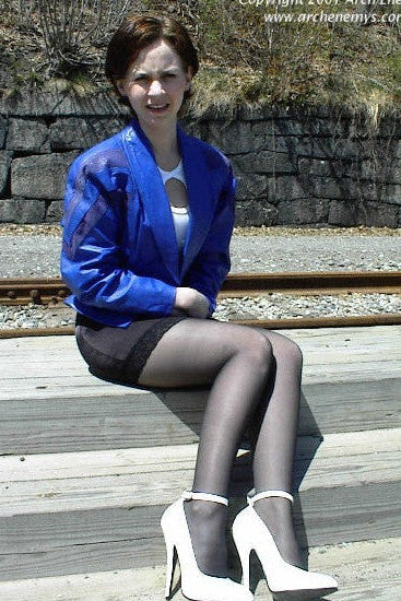 Woman in a blue jacket and black stockings sitting on a wooden platform with stone wall and tracks in the background.