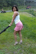 Woman in high heels pushing a lawn mower on a grassy area with a road and vehicle in the background.