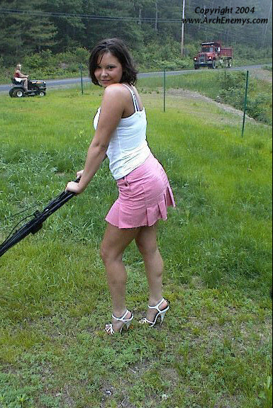 Woman in high heels pushing a lawn mower on a grassy area with a road and vehicle in the background.
