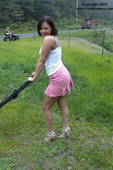 Woman in high heels pushing a lawn mower on a grassy area with a road and vehicle in the background.
