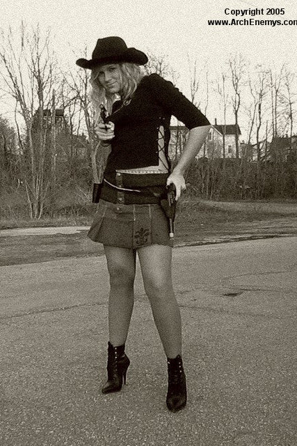 Person in a cowboy hat and skirt holding guns on a street.