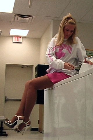 Woman sitting on a dryer in a laundromat wearing a white sweatshirt with a pink logo and a pink skirt and superarch heels