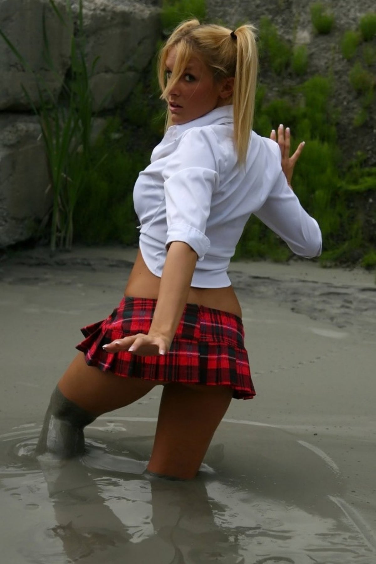 Woman in a white shirt and red plaid skirt standing in water near rocks and grass.