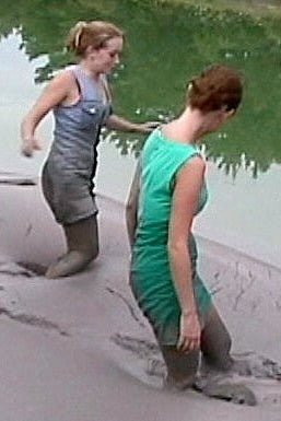 Two women standing in a shallow body of water, partially submerged in mud.