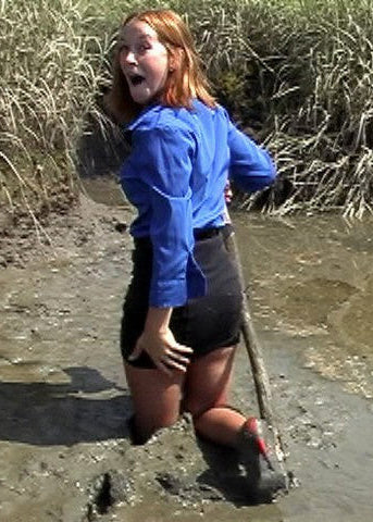 Person in a blue shirt and black shorts standing in a muddy area with reeds in the background
