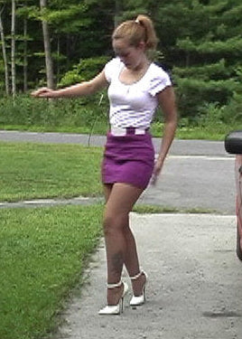 Woman in a white top and purple skirt standing on a sidewalk next to a red car.