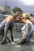 Two women covered in mud standing in a pool with concrete blocks and trees in the background.