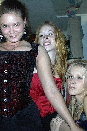 Three women posing together in a casual setting with shelves and bottles in the background.