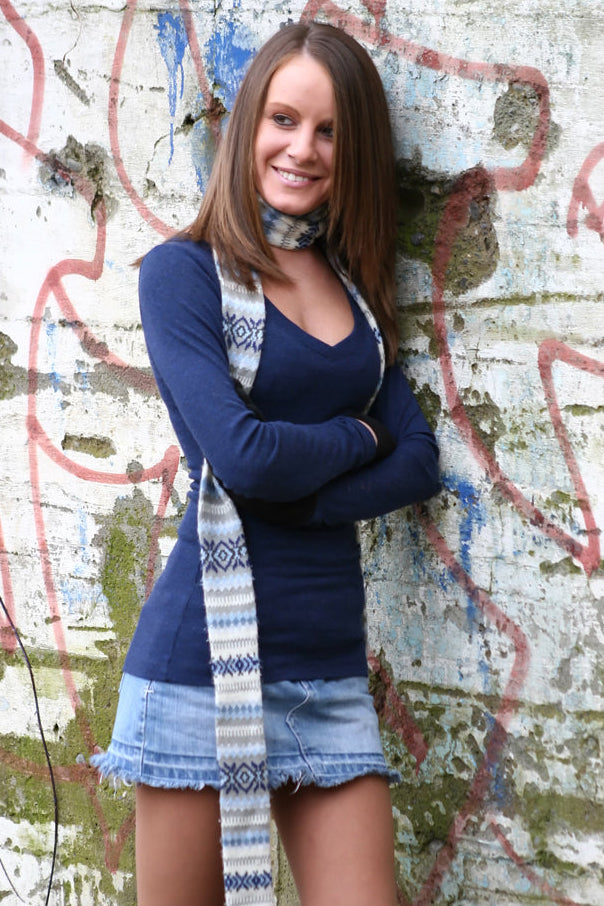A smiling young woman stands against a weathered graffiti wall, wearing a navy long-sleeve top, a light denim mini skirt, and a blue-and-white patterned scarf.