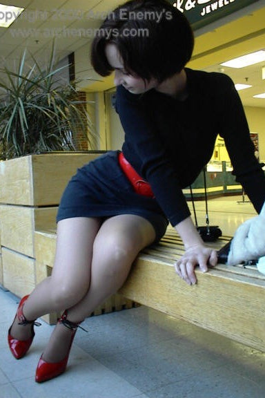 Woman in a black dress and red shoes sitting on a bench in an indoor setting.