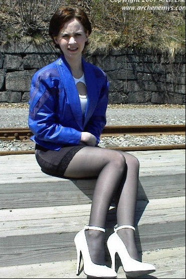 Woman in a blue jacket and black stockings sitting on a wooden platform with stone wall and tracks in the background.