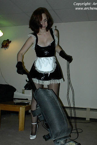 Person in a maid outfit holding a large black object in a room with a white wall.