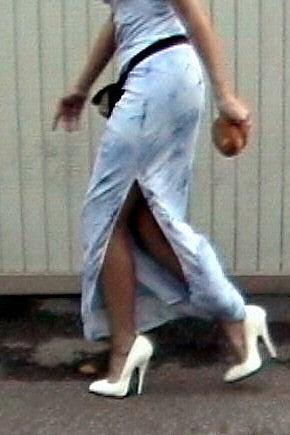 Person wearing a light blue dress with a high slit, white high heels, and a black belt. The background is a plain wall.